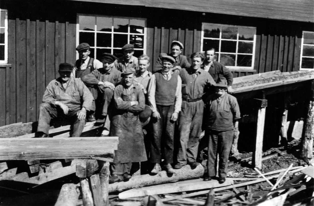 Gruppbild från Alingsås Snickerifabrik, kv Gjutaren. 11 män står/sitter framför en husvägg. Runt fötterna ligger bråte och olika träbitar.