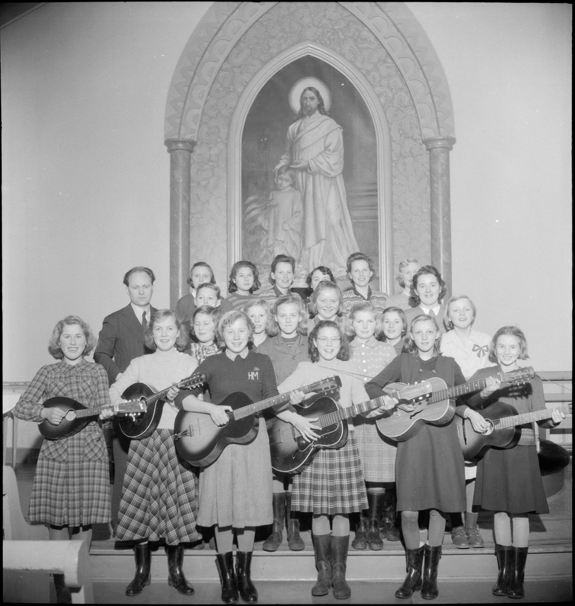 En  jentegruppe foran altertavlen i Metodistkirken. Flere av jentene har gitar eller ukulele.