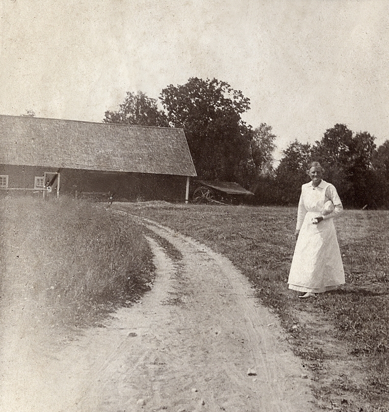 En kvinna i ljus klänning och förkläde står på en gräsplan, snett framför en ladugård.