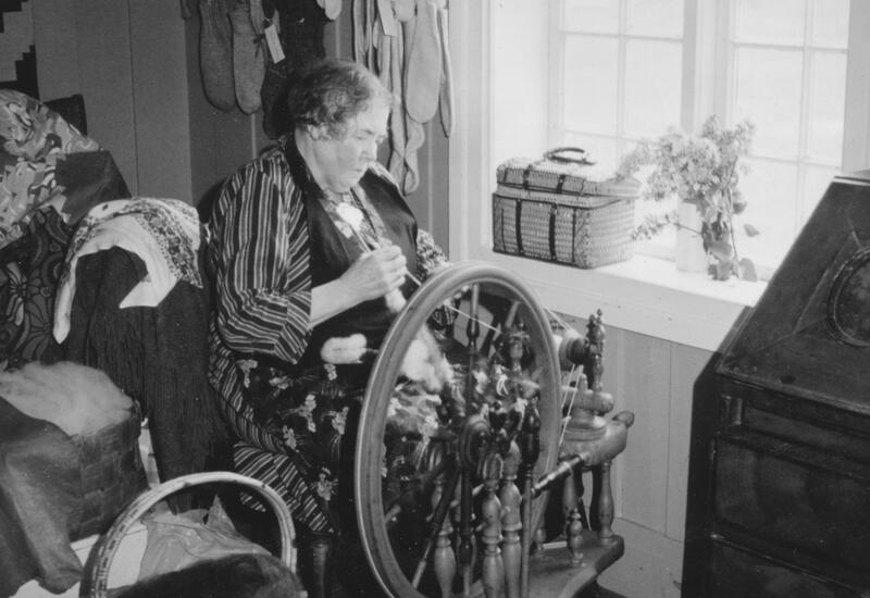 Foto i svart-hvitt av en dame som sitter ved en rokk. Fotografiet er med i nettutstillingen "Spansken".