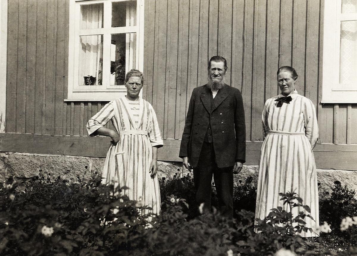 Två kvinnor i randiga klänningar och en man i kavajkostym står i en trädgård framför ett bostadshus.