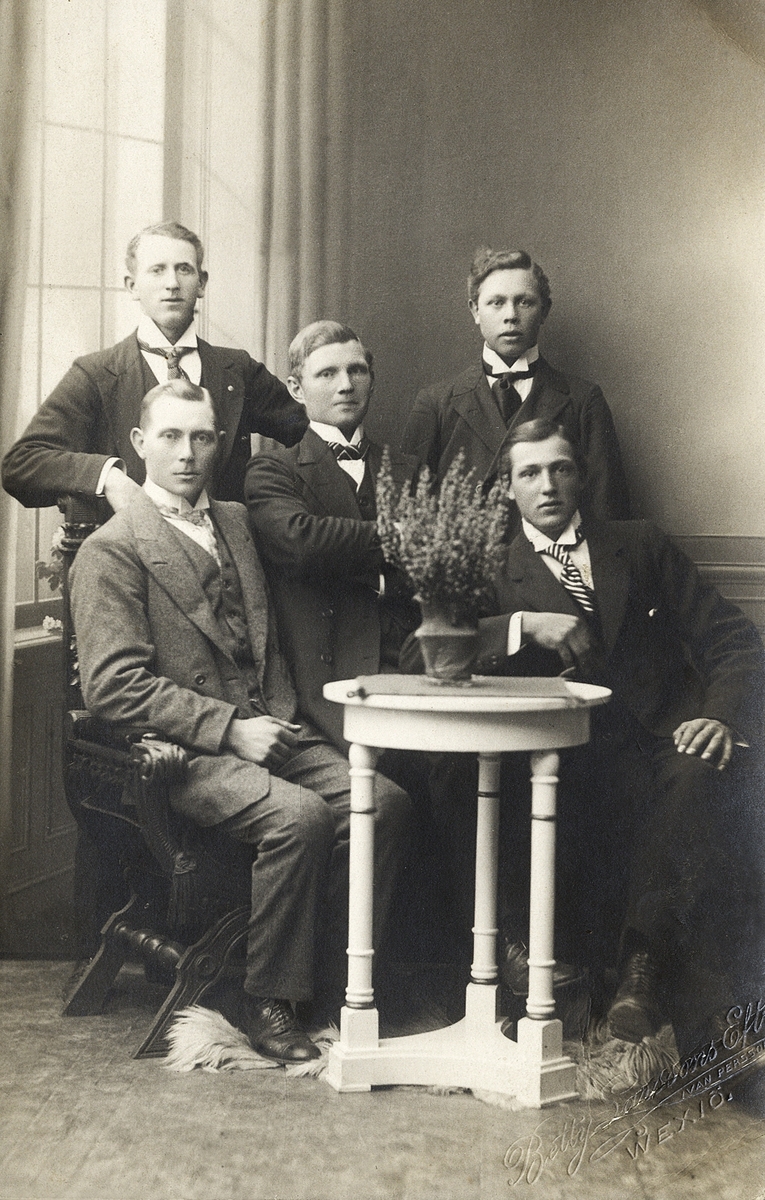 Fem unga män i kostym har samlats hos fotografen för ett gruppfoto. 
Tryckt som vykort, med fotografens stämpel i nedre högra hörnet.
På baksidan är skrivet männens namn: "Karl Svensson, Pehr Nilsson, Gustav Isaksson, Erik Johansson, Einar Gustavsson".