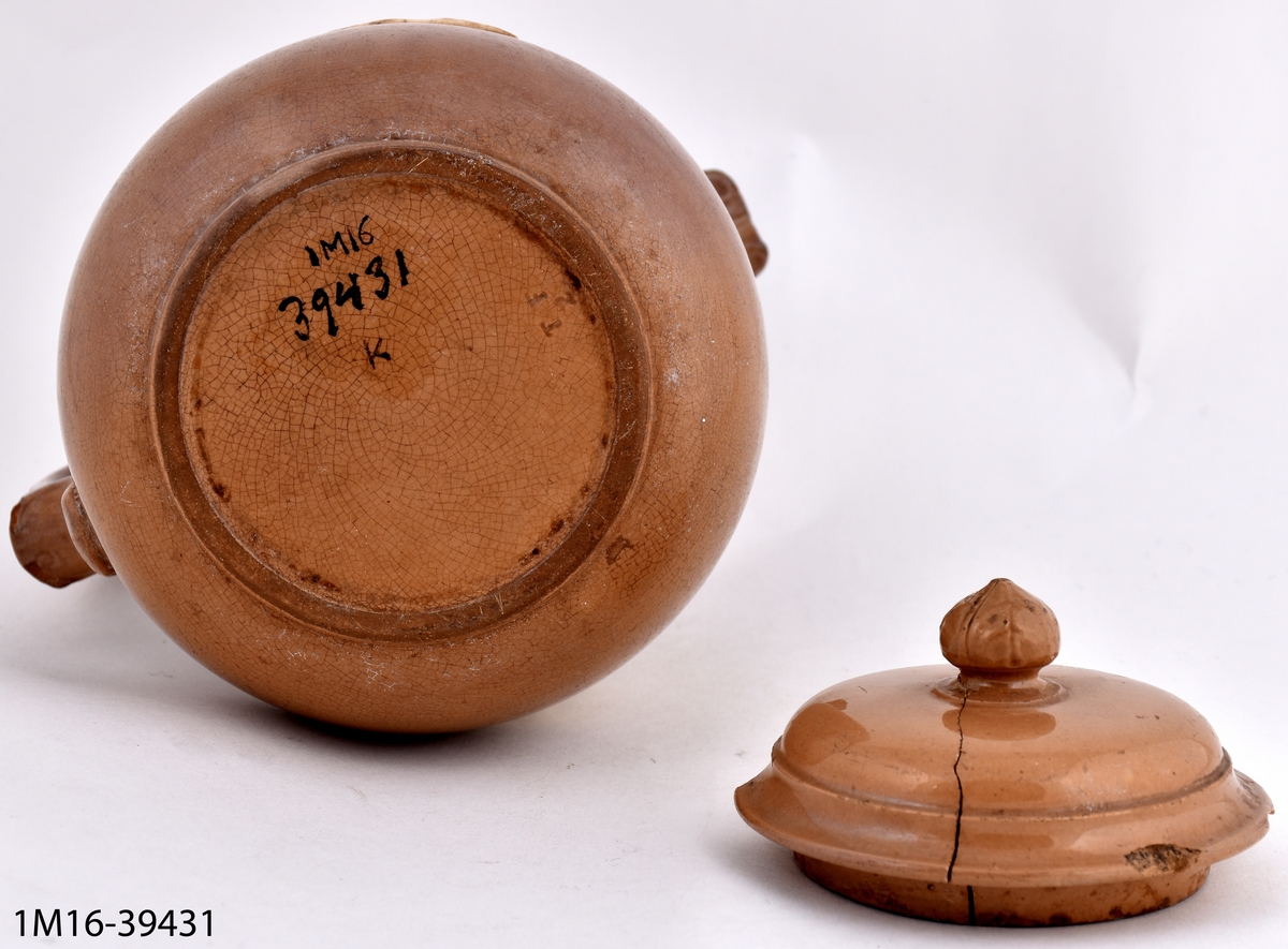 Tekanna, brun med 2 mytologiska grupper i vit högrelief på sidorna, ej pip men tut. Tillverkare: Tillinge Fayence och Terracotta Fabrik.