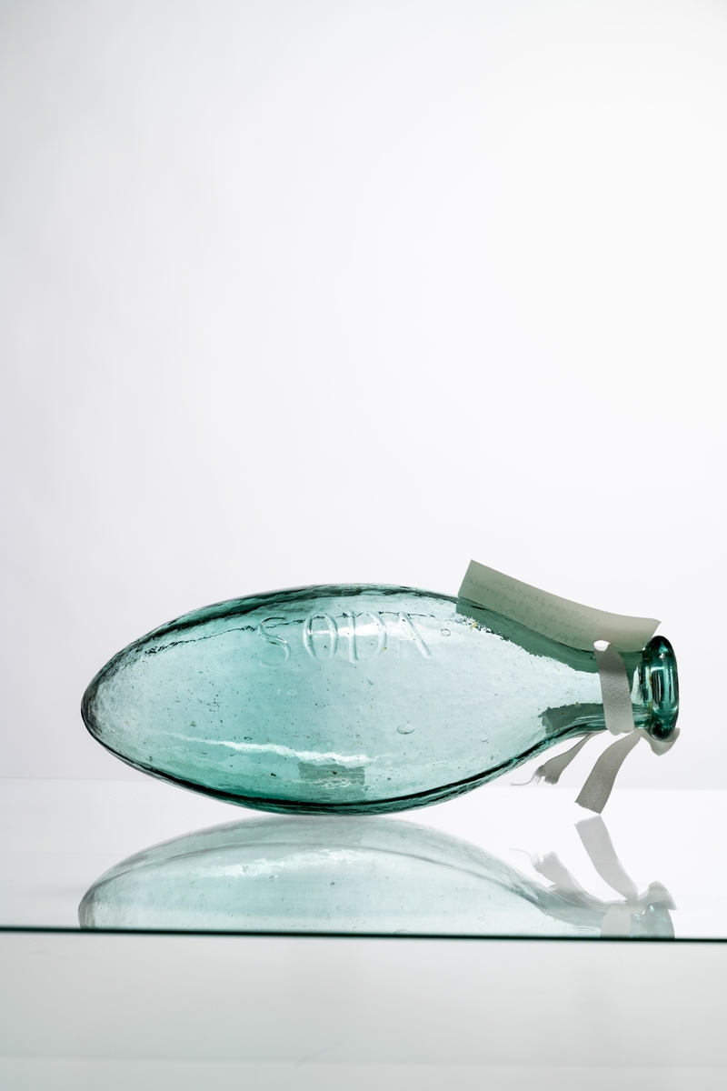 Flaska för vatten av ljust grönt glas, med oval, liggande form. Märkning i relief: "SODA". Flaskan gjordes med liggande form för att naturkorken inte skulle torka. Tillslutande kork saknas. Hantverksmässigt tillverkad vid Sunds glasbruk, Jönköpings län. Se vidare Historik.