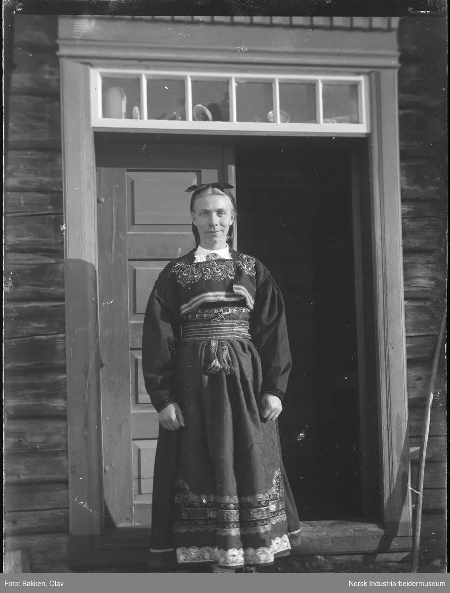 Portrett. Kvinne i bunad stående foran dør til bolighus. Overlysvindu over døren.