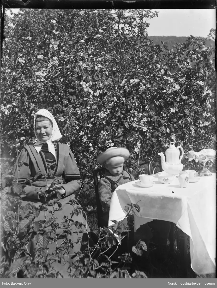 Kvinne og guttebarn sittende ved dekket bord i hage. Bordet har hvit duk og er dekket til kaffe.