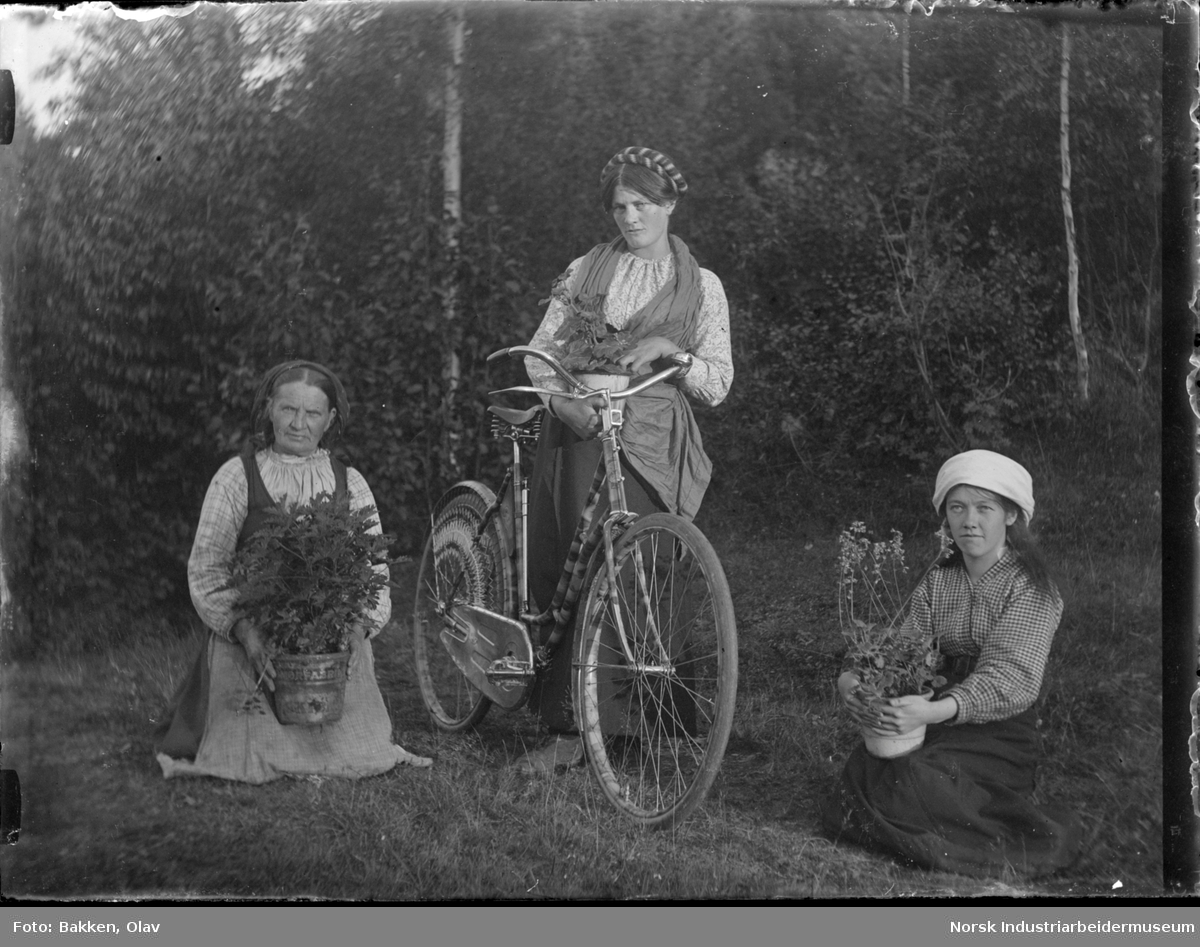 Tre jenter oppstilt utendørs. En med sykkel og potteplante i hånden. To sittende med potteplante på fanget.