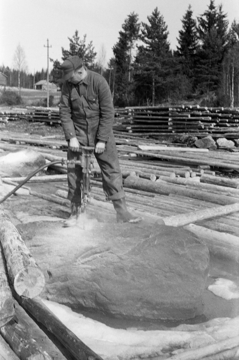 Steinboring med trykkluftbor på en velteplass ved Storsjøen i Nord-Odal i 1955. Mannen som utførte arbeidet var kledd i dongerikjeledress og hadde skyggelue på hodet. I de hullene han opparbeidet på denne måten skulle det legges sprengladninger som fikk steinblokken til å sprekke i mer håndterlige fragmenter, som lett kunne fjernes. Dermed skulle det bli enklere å rulle tømmeret fra strøveltene ut i vassdraget. Mannen arbeidet uten støvmaske, hørselvern eller annet verneutstyr.