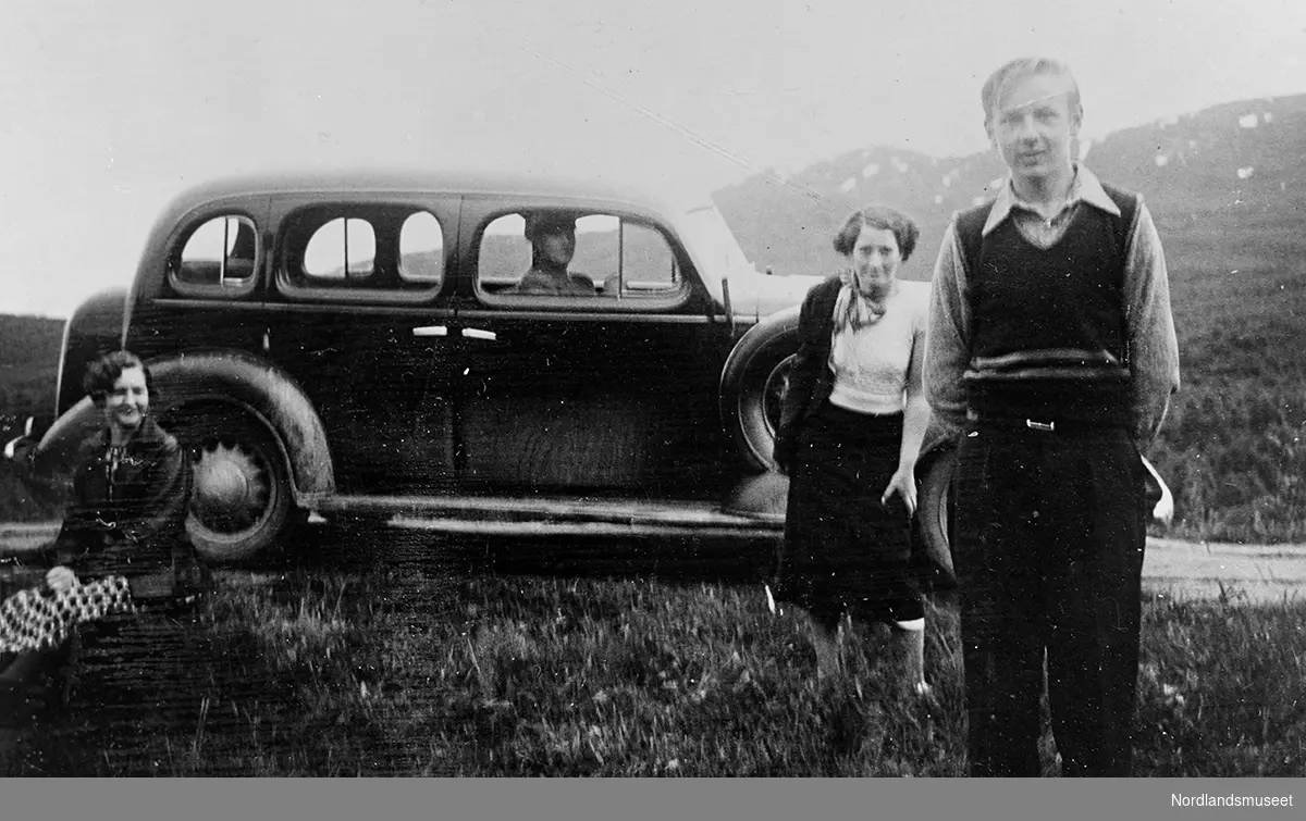 En bil, tre personer foran og en person sittende i bilen. Bildet viser Ballangens første drosjebil som var eid av Bjarne Sjåfjell som sitter i bilen. I forgrunnen helt til høyre er Johannes Sletteng, sittende ved bakhjulet på bilen er Marie Sletteng.