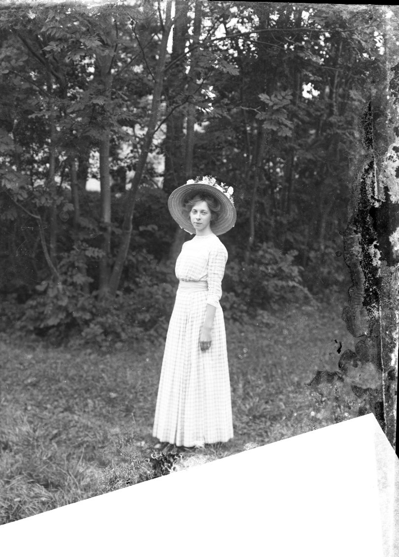 Porträtt av en ung kvinna i rutig klänning och hatt. Det är sommar. I bakgrunden anas Visingsborgs slottsruin.