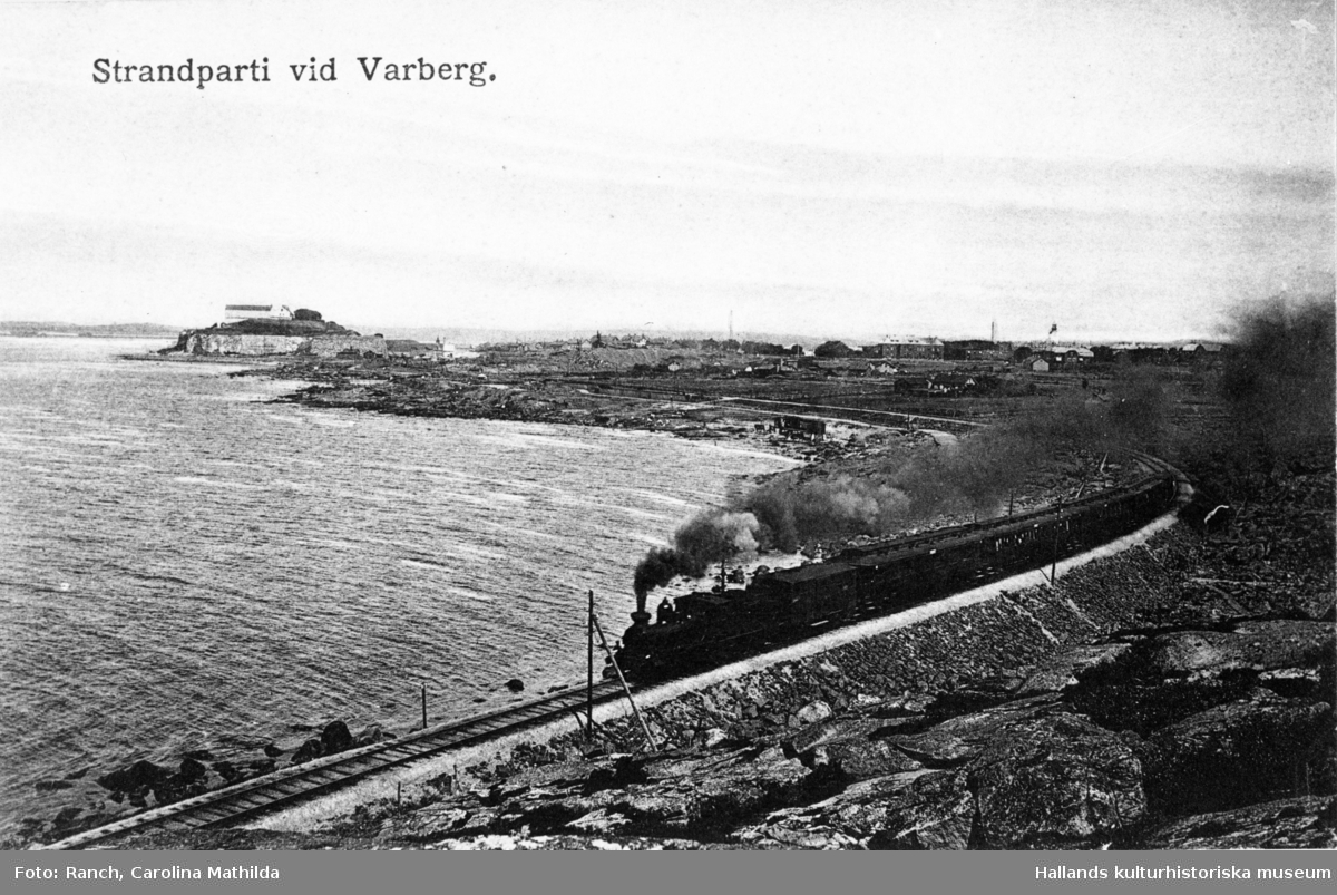 Vykort text: "Strandparti vid Varberg". Utsikt från Hästhagabergen över järnvägen söderut från Varberg, Mellersta Hallands järnväg, som invigdes 1886. Här kommer tåget ångande på järnvägsvallen längs havsranden. I bakgrunden syns Varbergs fästning.