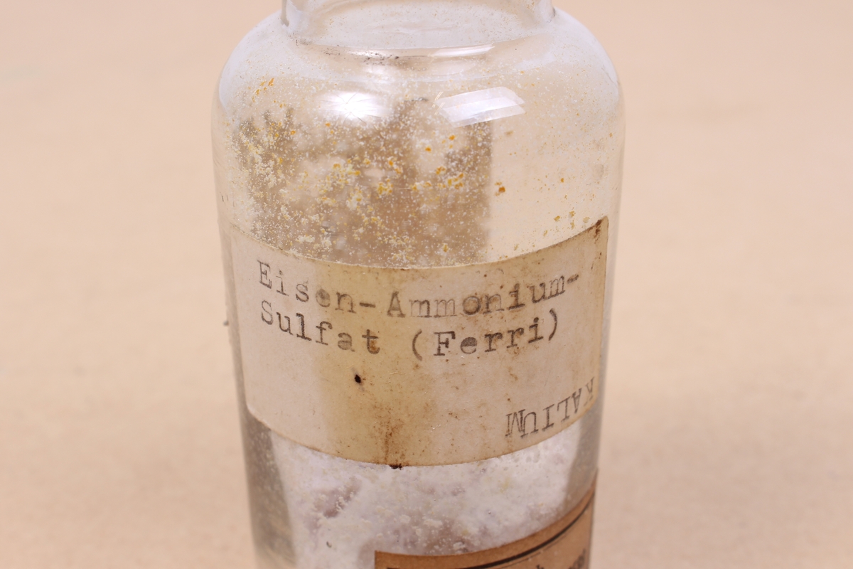 Kjemikalieflaske av klart glass, med stopper av kork. Inneholder ammonium jern(III)-sulfat, en type alunsalt som har blitt brukt blant annet i garving.