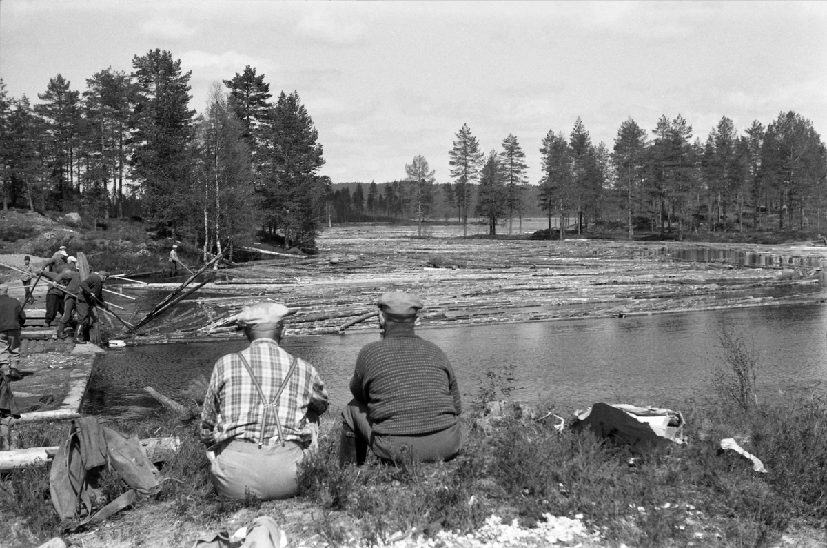Tapping av tømmer fra Nøklevatnet, som ligger i gresesona mellom kommunene Nord-Odal, Grue og Åsnes i Hedmark. Dette fotografiet fra 1959 skal være tatt under den første fløtinga fra denne innsjøen siden cirka 1920. De to karene som satt i forgrunnen med ryggen mot fotografen skal ha vært veteranene Rolf Bakken (1897-1960) og Menz Berg (1910-1998). De satt med blikket vendt mot en lukedam der det ble tappet vann og tømmer. Ei gruppe fløtere befant seg på dambrua, der de, ved hjelp av fløterhaker, forsøkte å trekke fløtingsvirket riktig inn i damløpet.