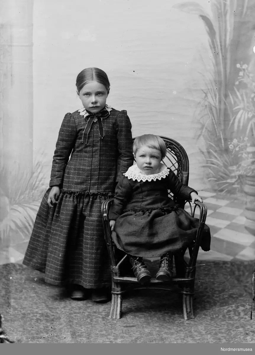 To barn, en jente stående ved stol. En enda mindre jente sittende i stolen. Atelierbakgrun med søyler og palmer.