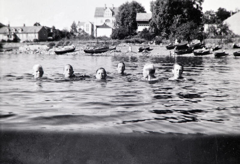 Svart-hvitt foto av seks mannshoder som stikker opp av vannet under en svømmetur. Bymiljø i bakgrunnen. Koigen, Hamar. (Foto/Photo)