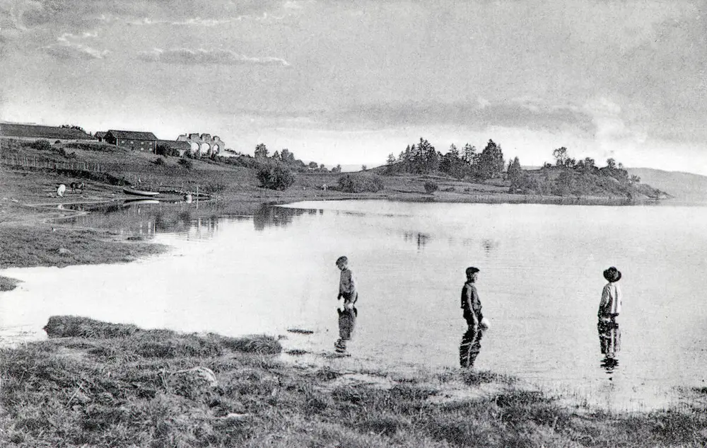 Svart-hvitt fotografi av tre barn som vasser i Mjøsa i Rosenlundvika, domkirkeruinen vises i bakgrunnen.