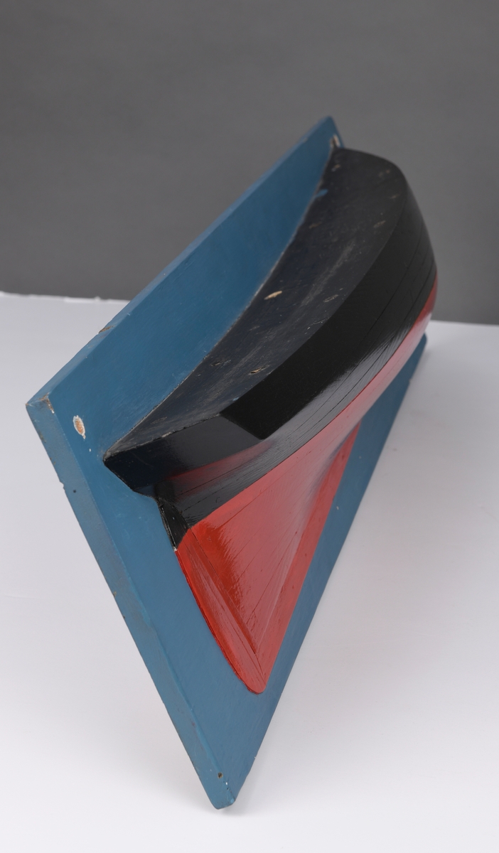 Halvmodell/ skivemodell av sluppen "Vidar". Gjennomgående skruehull for veggmontering på den blå bakplaten. Modellen er laget av tre, og er malt i sort og rødt.