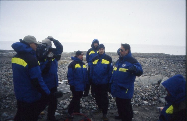 Ett antal personer i polarforskningssekretariates blågula plagg står på Kvitöya (Vitön). En man talar till gruppen. Till höger står en annan man och filmar talaren.