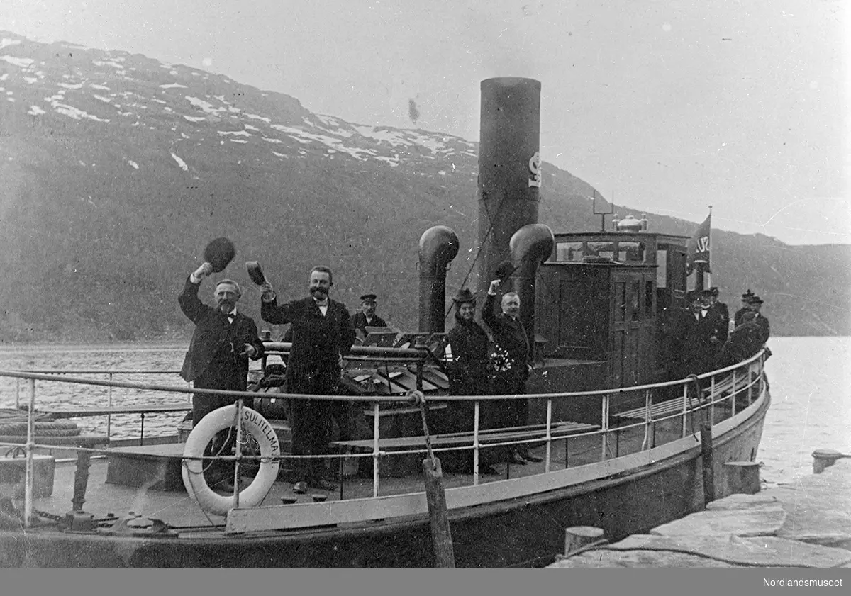 Ombord i slepebåten "Sulitjelma III" ved kaia i Furulund. Båten ligger utenfor "Arne Jørgensen-butikken". Bildet viser mennesker som vinker fra dekket på båten.