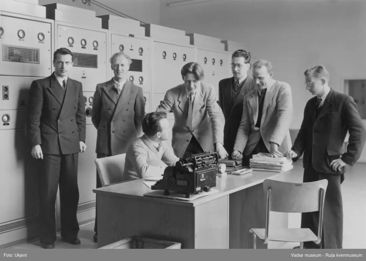Et teknisk rom inne i NRK-bygget i Vadsø. Vi ser 6 menn  kledd i dress, skjorte og slips, står rundt et skrivebord. Ved skrivebordet sitter en mann kledd i v-genser, skjorte og slips. På skrivebordet en del dokumenter og en skrivemaskin. Bak mennene en hel vegg dekket av teknisk utstyr.  
Finnmark kringkaster (NRK-bygget) i Vadsø, byggeår 1949. Arkitekt Henrik L. Kjær. Bygget har bærende betongvegger og fasadene er lagt med teglstein og grunnmuren er belagt med skiferplater. (Kilde: Arkitekturguide for Nord-Norge og Svalbard)