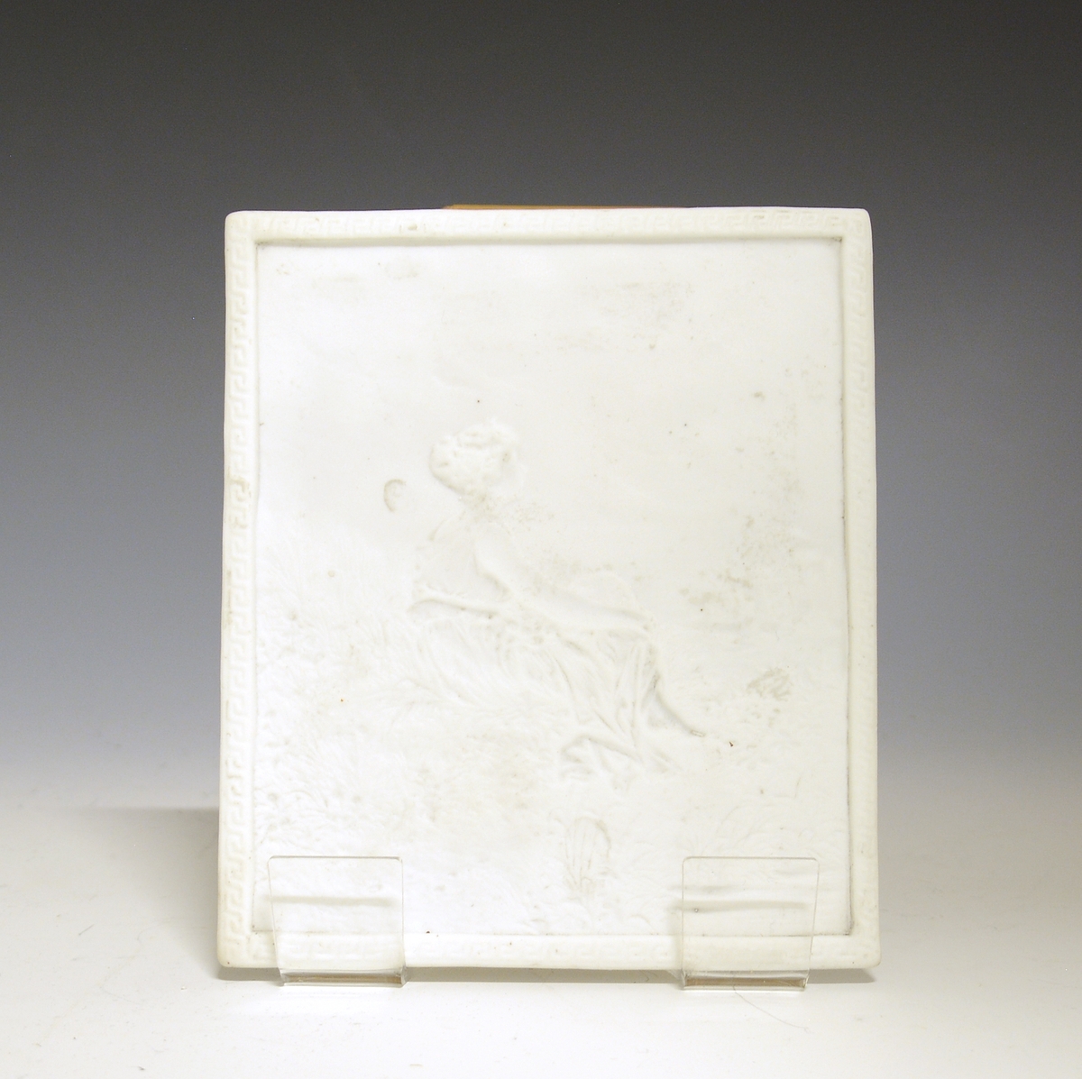 Prot: "Lysbilde" - tynn plate av porselen med smal ramme som har à la greque-bord. Motivet, en sittende kvinne i landskap, er delvis utført i bas-relieff. Uten glasur, uten mrk. og sign.
