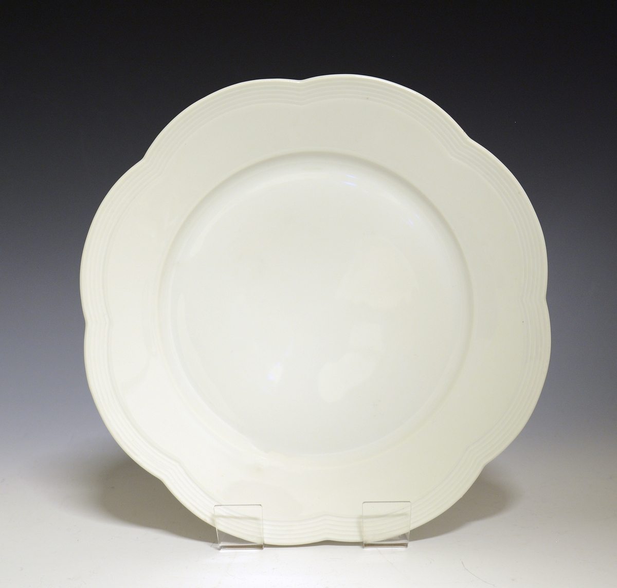 Prot: Flat tallerken av porselen. Bølget kan som er markert med et bånd av fine riller som følger den bølgede kanten,