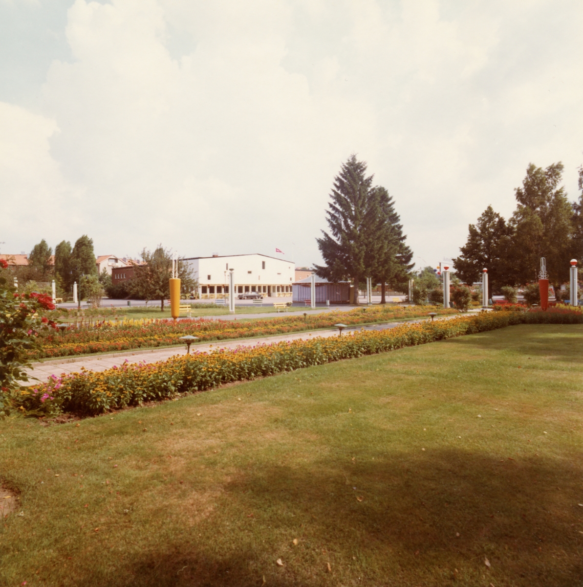 Bild från parken i Folkets Park. I bakgrunden en stor vit byggnad. I förgrunden en gräsmatta och en blomrabatt längs med gången.