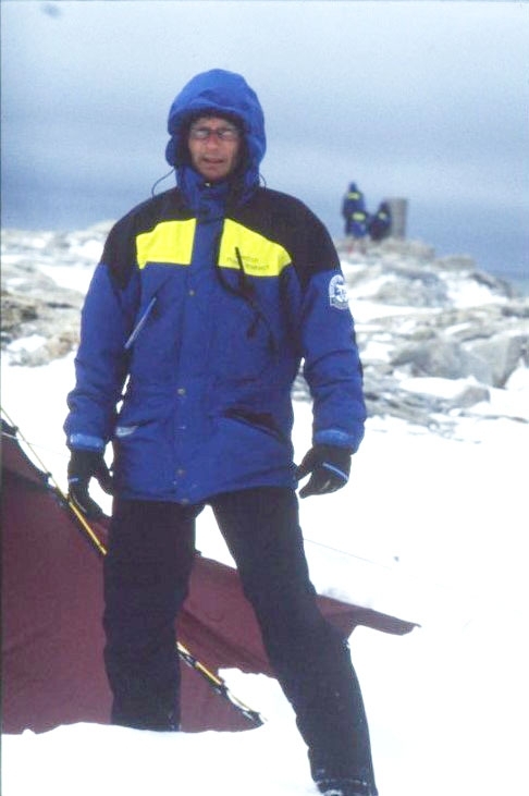 Porträtt av en man med glasögon, i polarforskningssekretariatets klädsel. Han står vid ett tält vid Andréelägret på Vitön med huvan uppfälld, det blåser och är kallt.