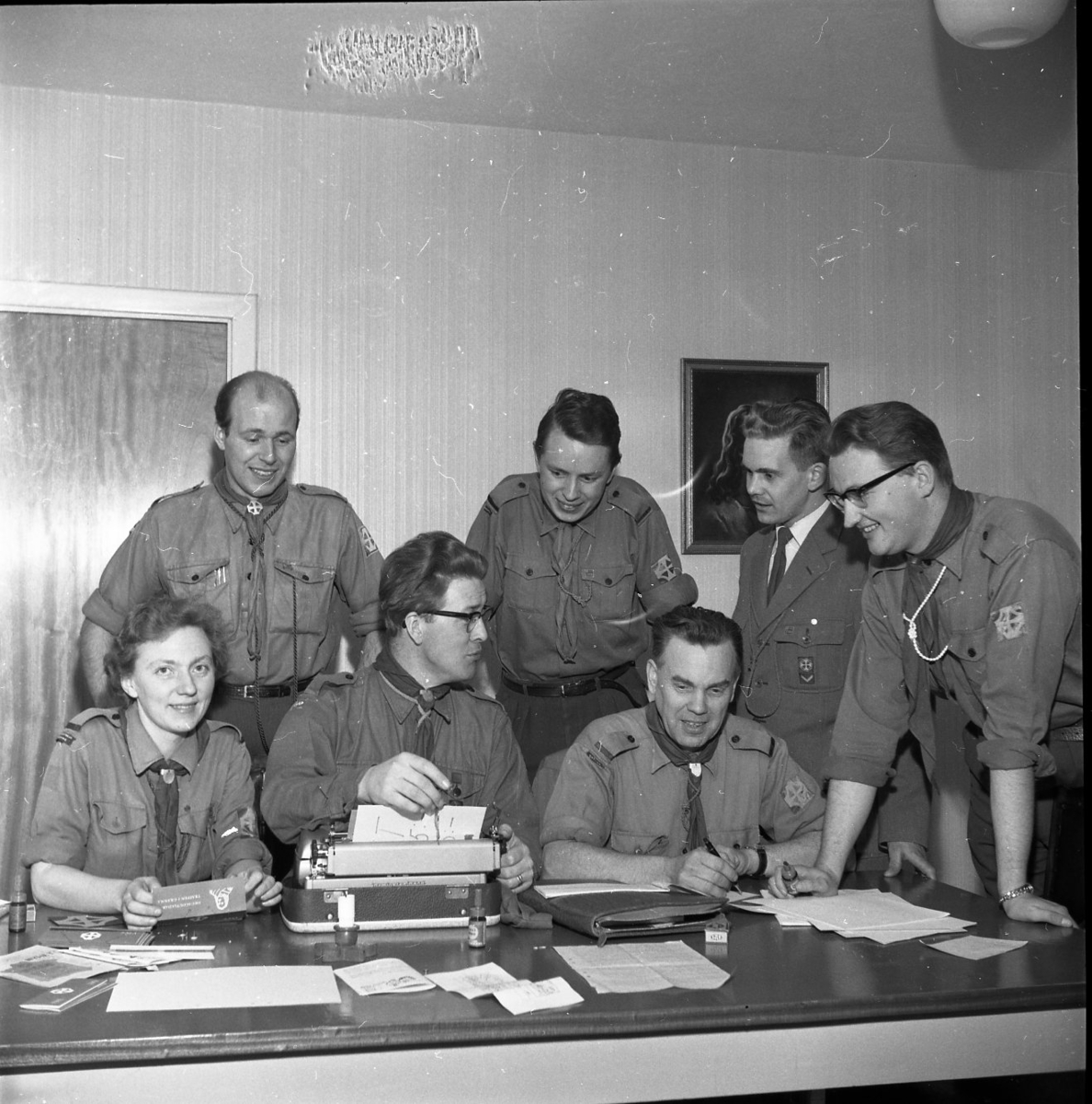 Sex män och en kvinna befinner sig vid ett skribord. Tre personer sitter, varav en sitter framför en skrivmaskin. Fyra personer står lutade över de sittades.