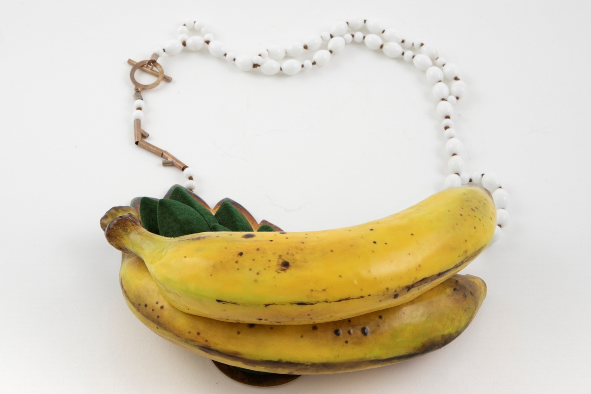 Skulpturell halssmykke med to naturtro bananer i gummi og blader av pålimt grønn flokk (minner om velur), festet på en sølvplate. Kjede med hvite glassperler i ulik størrelse og enkel lås i sølv.