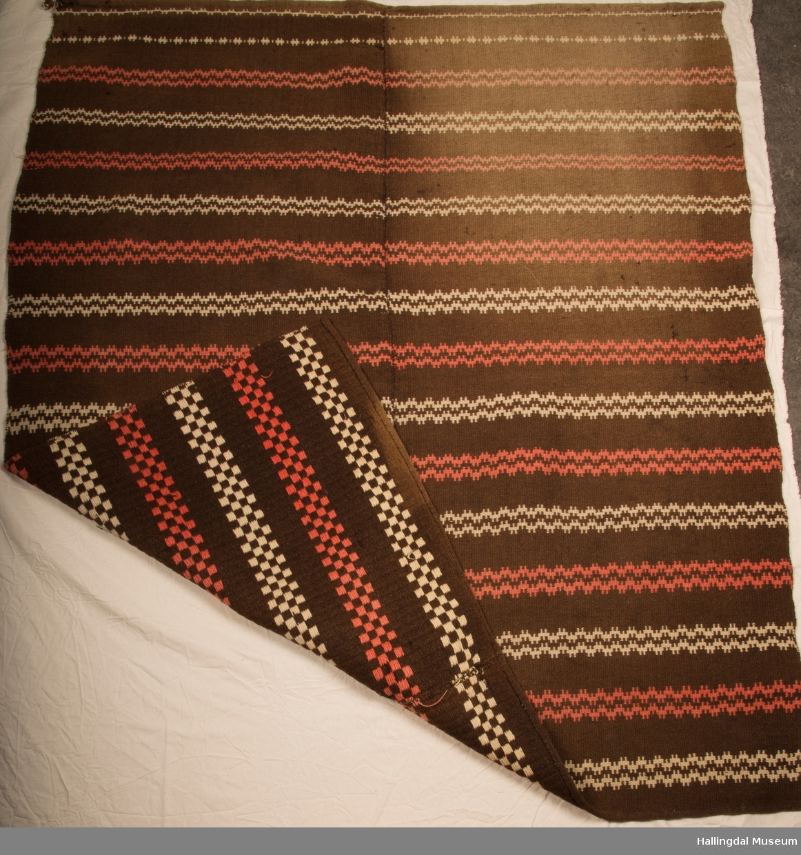 Åkle, sengeteppe som er sydd sammen av to høyder.  Vevd i krokbragd, brun bunn med hvite og røde sikksakk border.  Håndsydd.  Renning av bomull og ull innslag.