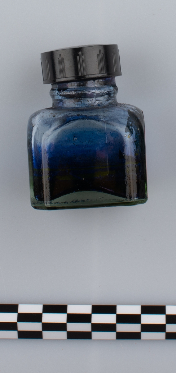 Glassflaske med tørkede rester av formentlig blått blekk. Sort skrulokk i kunstmateriale. Etikett på en side.