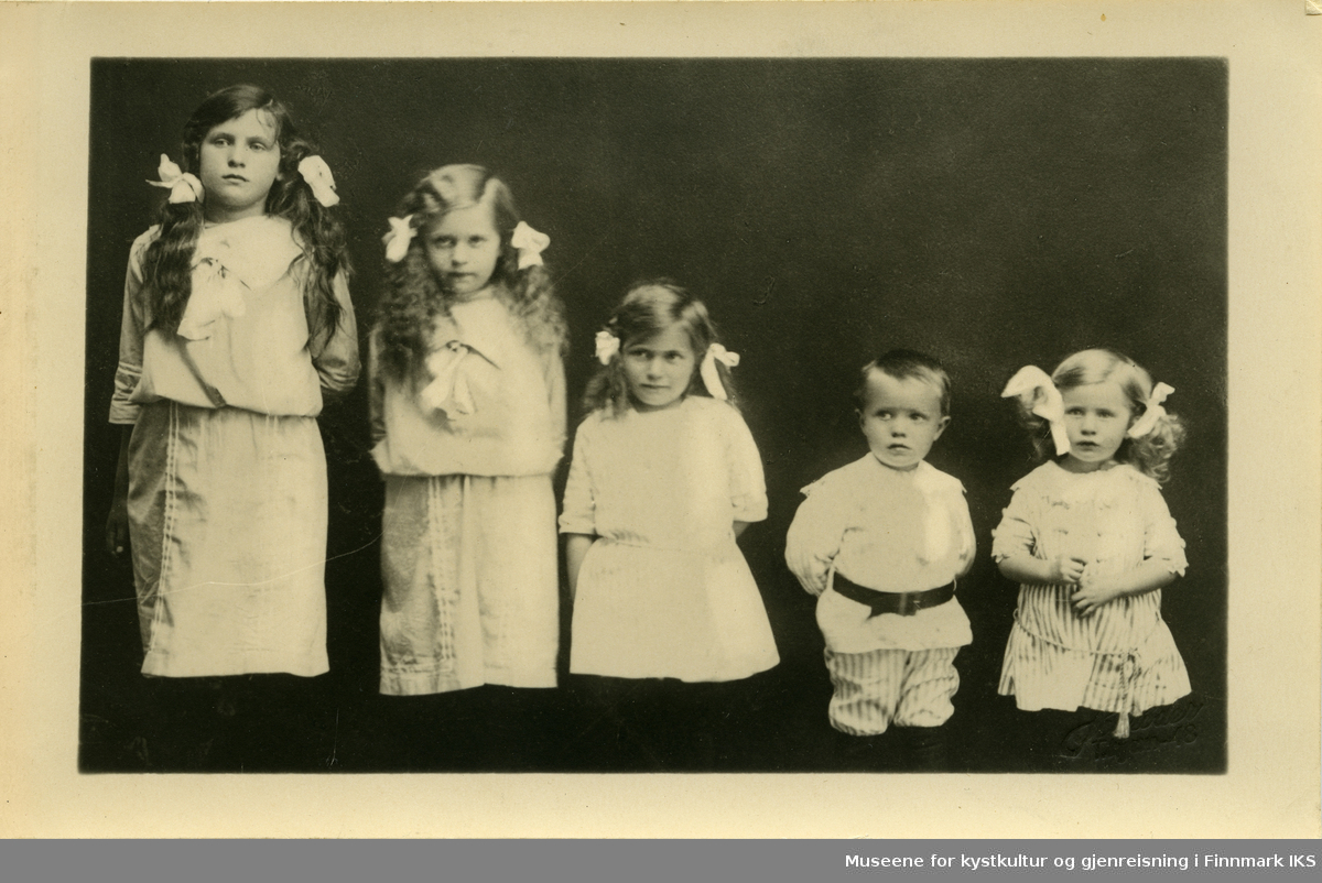 Fem barn har stilt seg opp i sine lyse finklær hos fotografen. Jentene har musefletter med sløyfer.