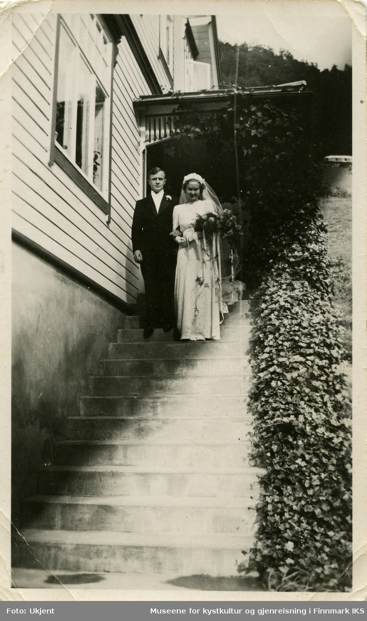 Et nygift par står på en trapp. Han har på seg en mørk dress med en blomst festet til, hun har på seg en brudekjole og holder en blomsterbukett.