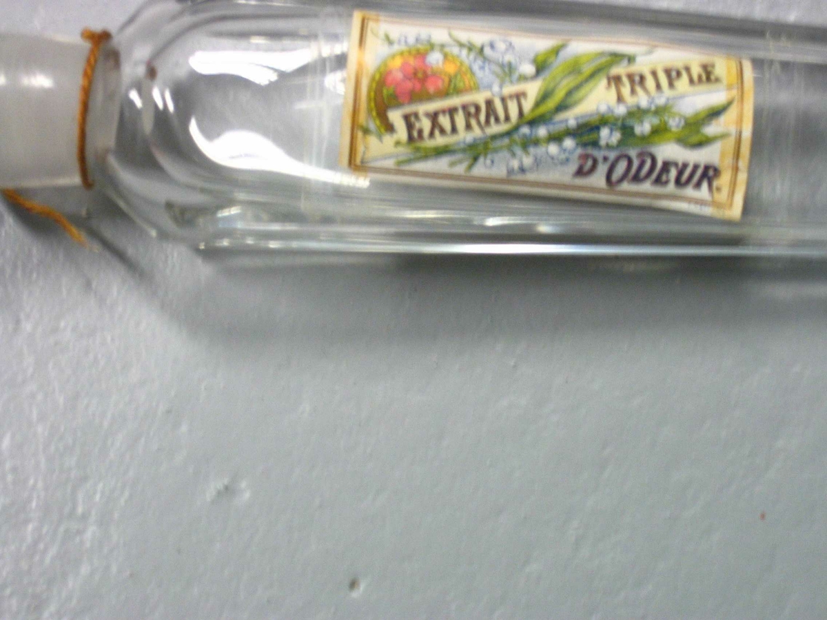 Avlang parmymeflaske av glass med propp. Pålimt papiretikett med tegning av liljekonvalller og andre blomster og teksten EXTRAIT TRIPLE D'ODEOUR