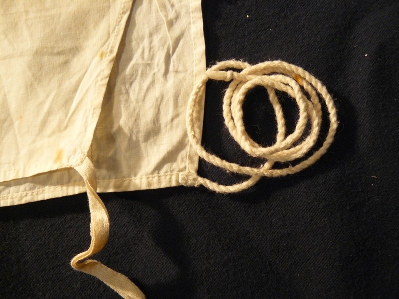 Kjolekrage av hvitt stivet bomullsstoff med blondekant. Har skulder og forstykke med snorer. Tvinnet bomullsnorer nedre kant på bakstykket og bomullbåndhemper i forstykkene.