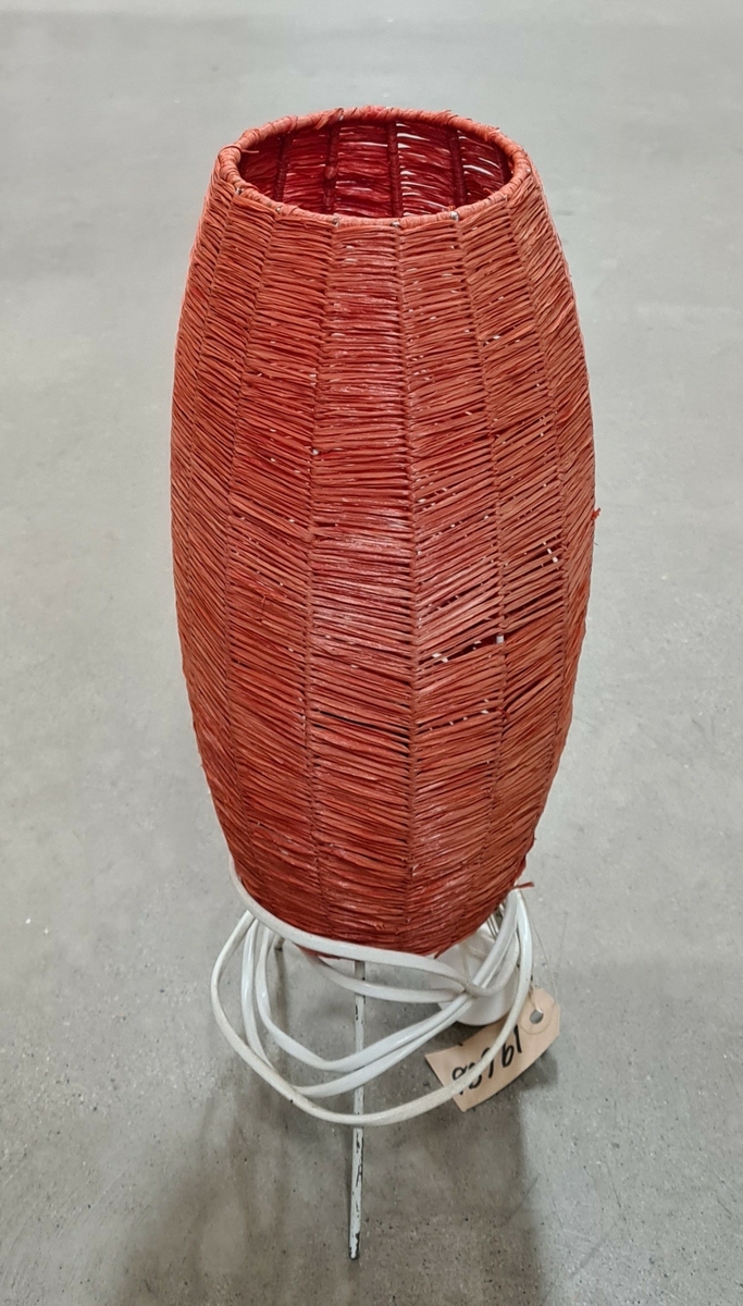 Bordslampa. Röd skärm av flätad bast, tre fötter, vit sladd med strömbrytare.

Inköpt 1985 till utställning om 1950-talet producerad av Älvsborgs länsmuseum.