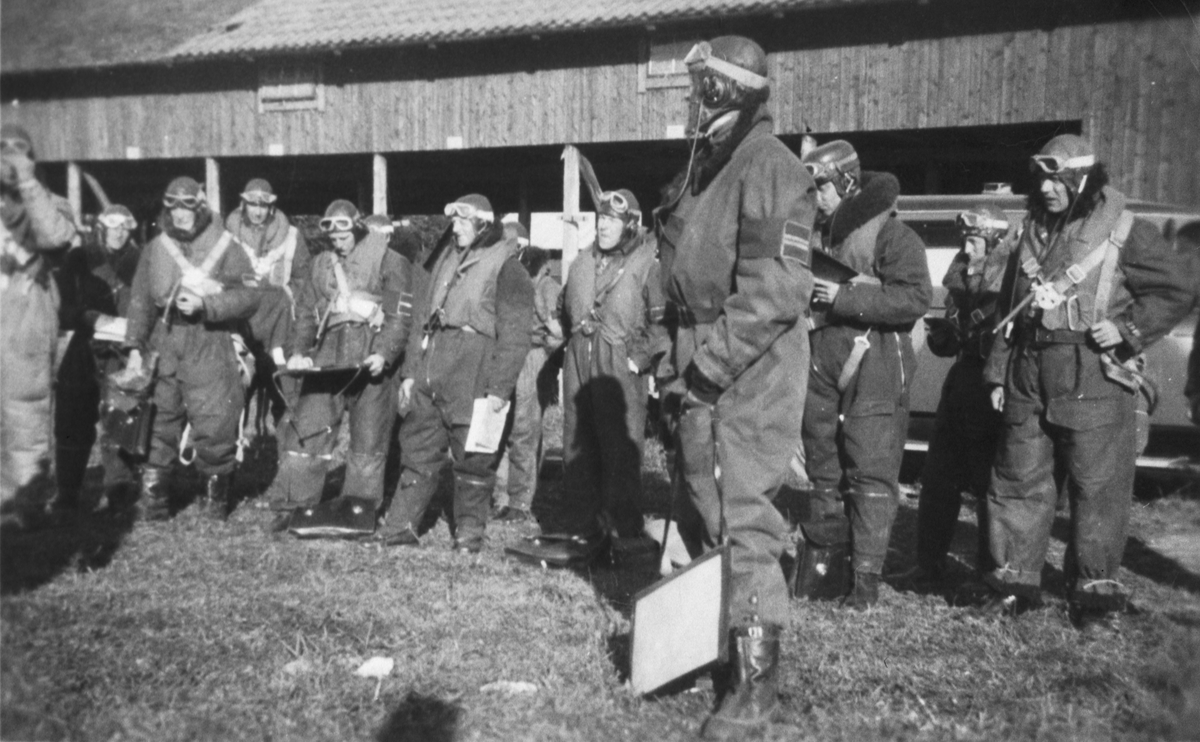 Tretton militära flygare får genomgång framför en lada innan flygning.
Troligen manöver vid Tierp, 1925.