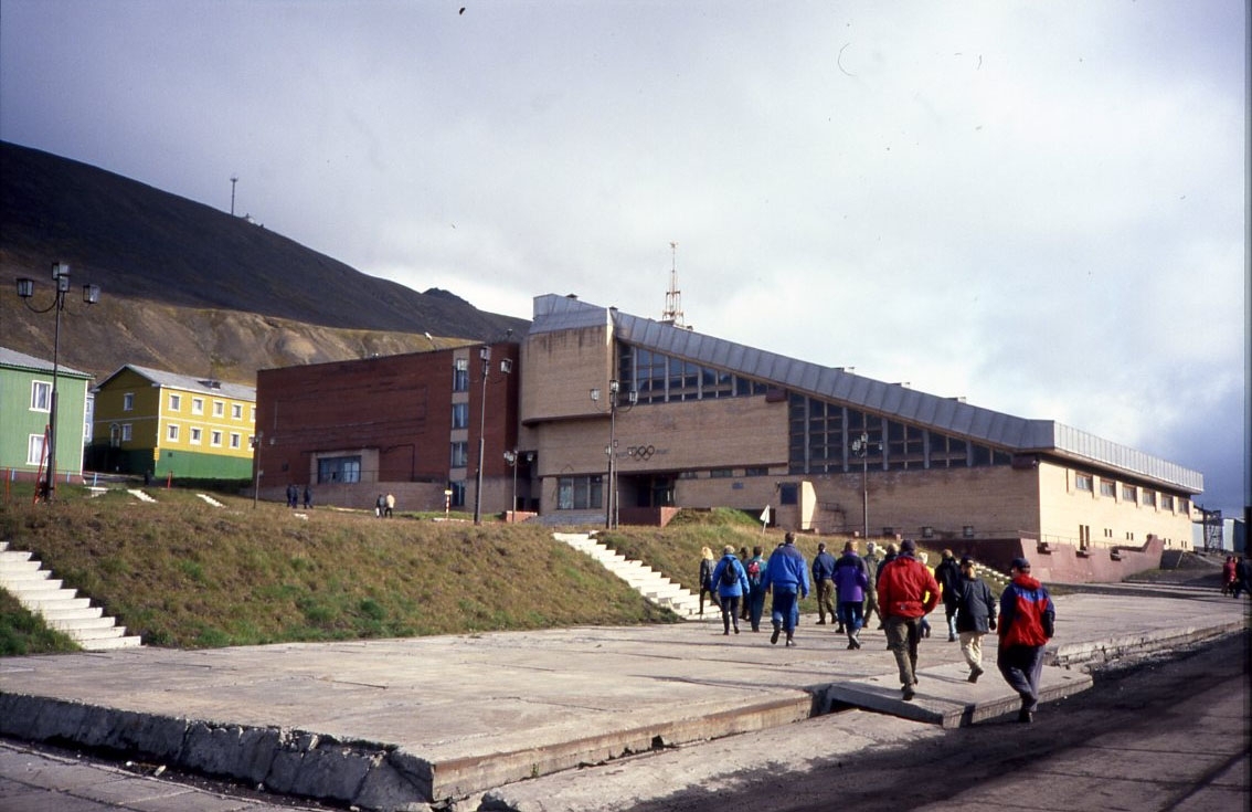 Bebyggelse i det ryska gruvsamhället Barentsburg på Svalbard. En grupp turister rör sig upp mot några byggnader. Till höger idrottshallen med OS-ringarna på väggen.