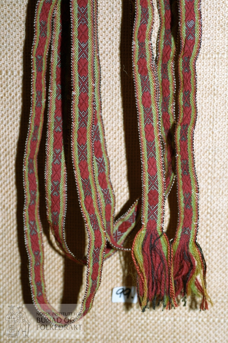 Plukkaband med renning av tynn svart silketråd og gulgrønt, grønt og rødbrunt mjukullsgarn. Innslag av kvit silketråd. Frynser av renninga i endene. Bredde: 1,5 cm. Lengde: 240 cm.