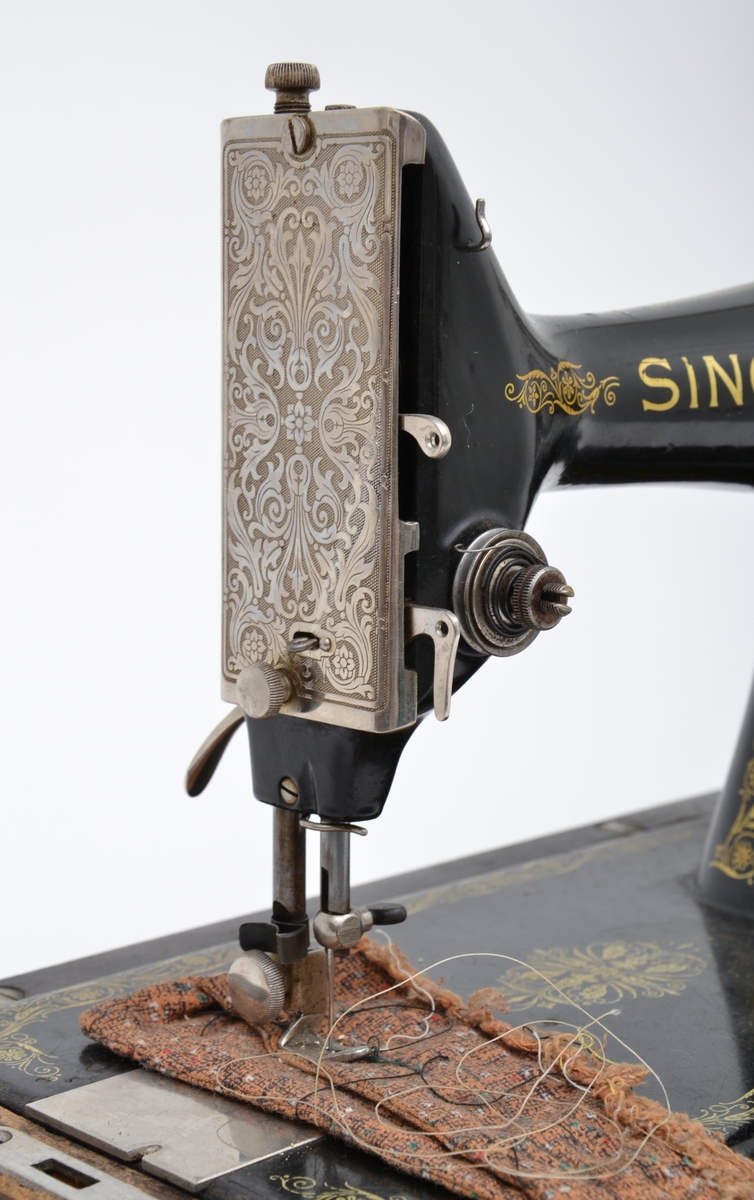 Manuell Singer symaskin med rankemotiv dekorasjon, lokket er laget i tre og det er bueformet med påklinte lister. Kiler på innsden. Håndtaket er skrudd på.


