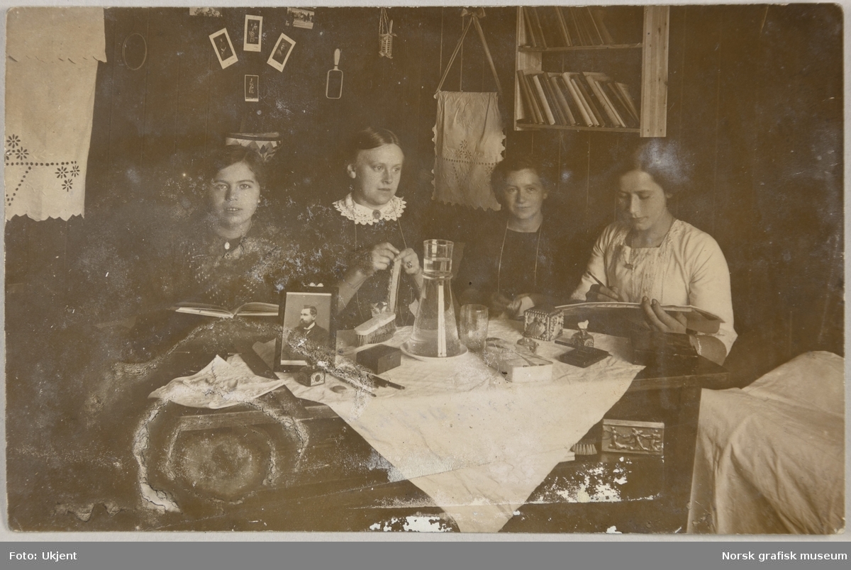 Postkort med bilde av fire kvinner. De sitter rundt et bord og leser, hekler, syr og tegner. På bordet står et bilde av en mann. Det er også børster, penner og blyanter, blekk, en vannkaraffel og et skrin.
