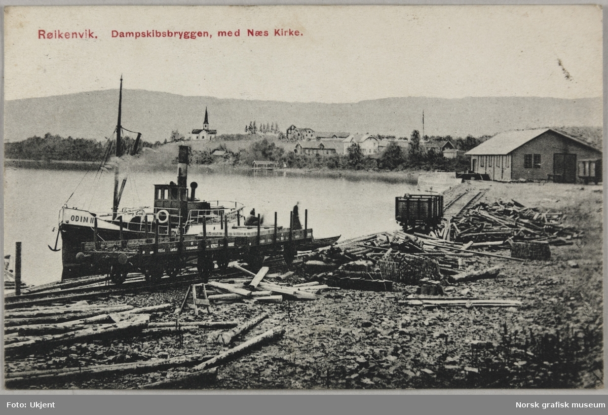 Postkort med bilde fra Røykenvik ved Randsfjorden. Det ligger en liten dampbåt ved kai, med trelast rett ved sjøkanten. I bakgrunnen er Nes kirke.