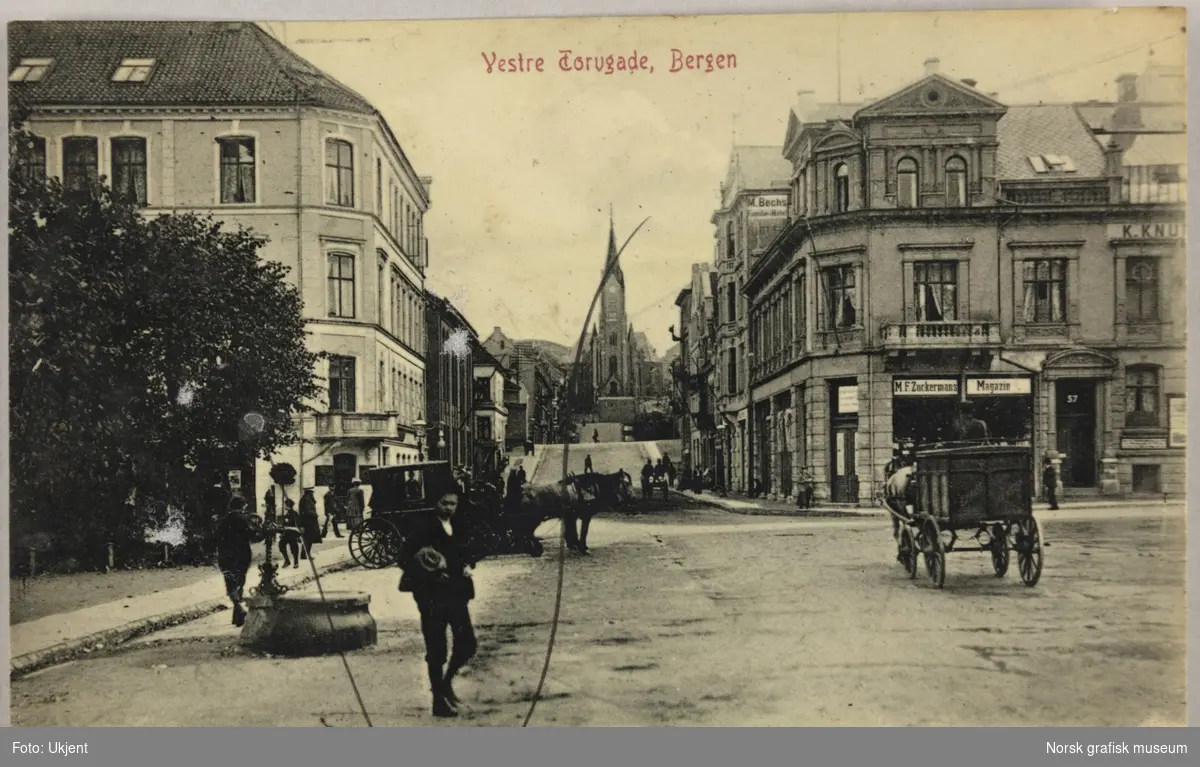 Postkort fra Vestre Torggaten i Bergen. Det er folk og hestevogner i gata. I bakgrunnen ligger Johanneskirken.