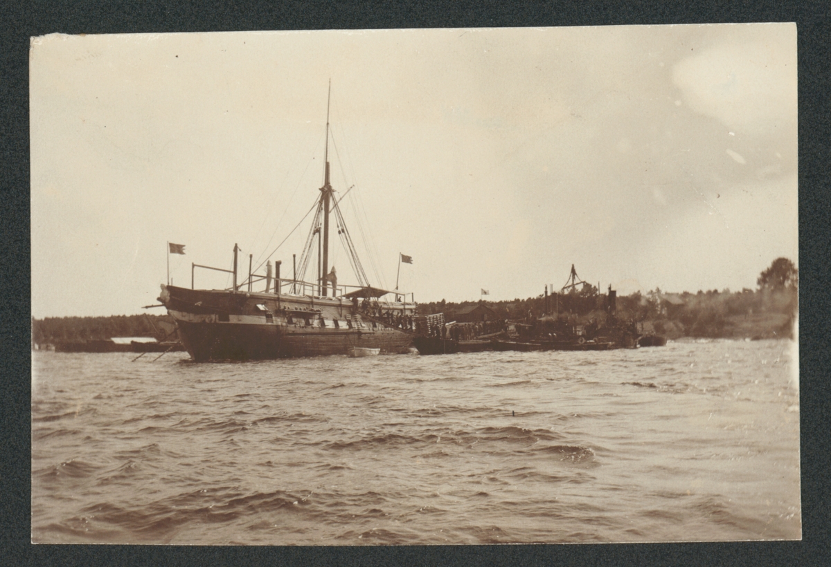 Bilden visar fregatten Af Chapman nedriggad efter ombyggnad till logementsfartyg. Vid sidan ligger flera små båtar förtöjd. En grupp soldater lämna fartyget genom landgången. I bakgrunden ser man en midsommarstång.