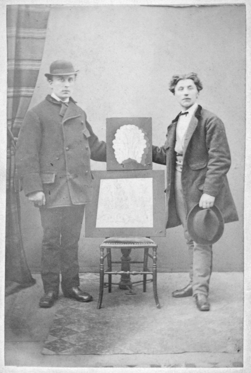 Et visittkort med et portrett av maler Alex Andersen (til venstre) og en venn. De står med to malerier/kunstverk mellom seg, støttet oppå en stol. På baksiden av visittkortet står det "Mit Kunstværk er det nederststaaende. A. Andersen".