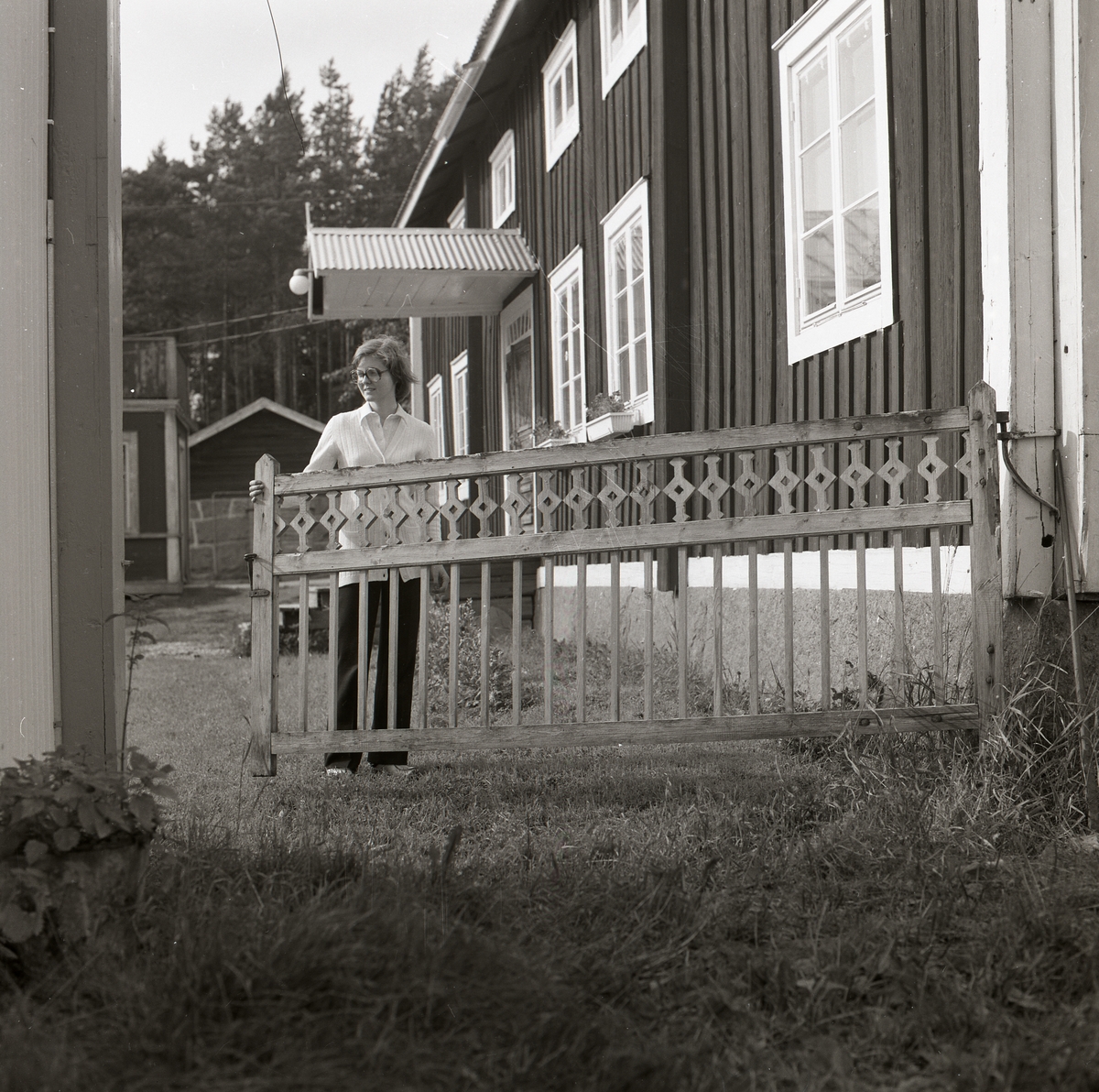 En kvinna öppnar en trägrind med spjälor som sitter mellan två hus, Trönö 1978.