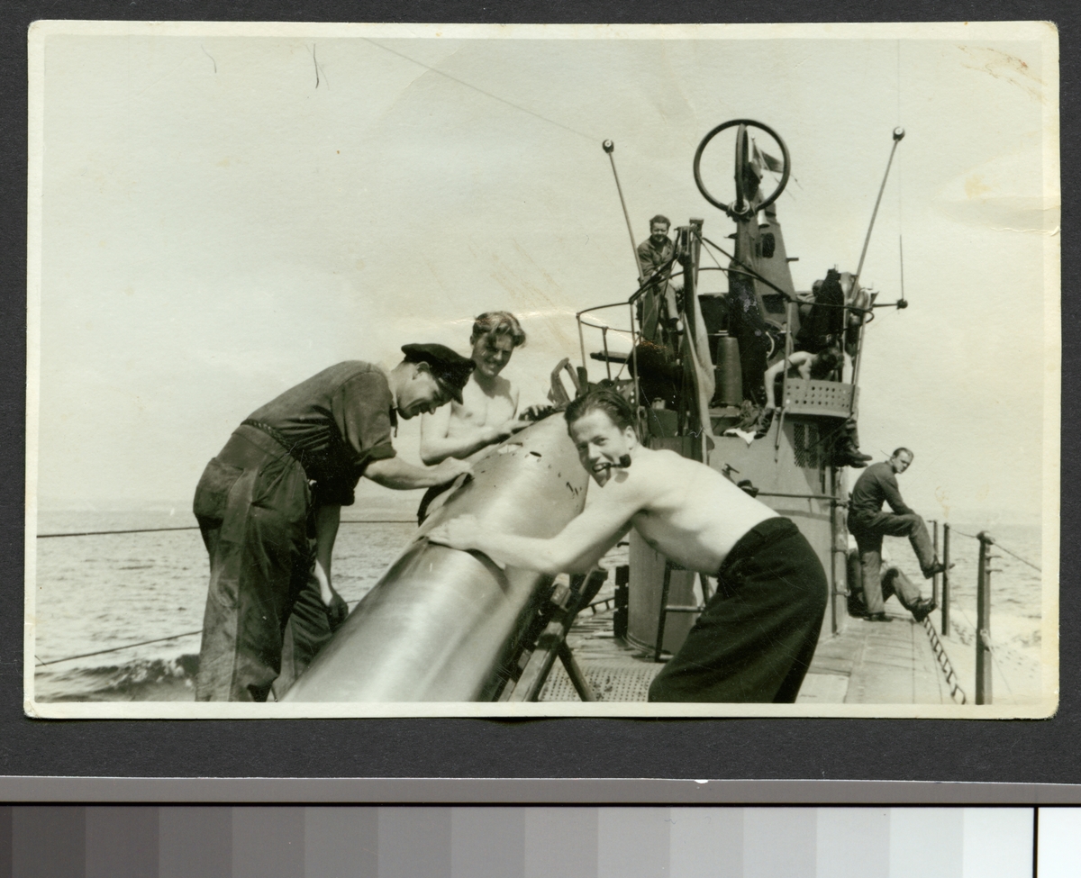Bilden visar tre sjömän som arbetar med en torped på däck av ubåten Najad. En man bär arbetskläder de två andra arbetar med bar överkropp och mannen in förgrund röker en pipa.
