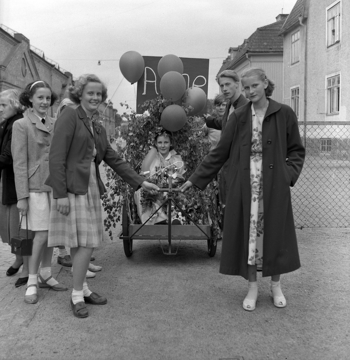 Ett antal personer kör en lövad vagn med en vän som tagit realen eller studenten. Från vänster: okänd, Anita Olofsson, okänd, Elisabeth Larsson (gift Danås), Anne (i vagnen), okänd, Bo Reteike, Rut (syster till Anne). Juni 1953