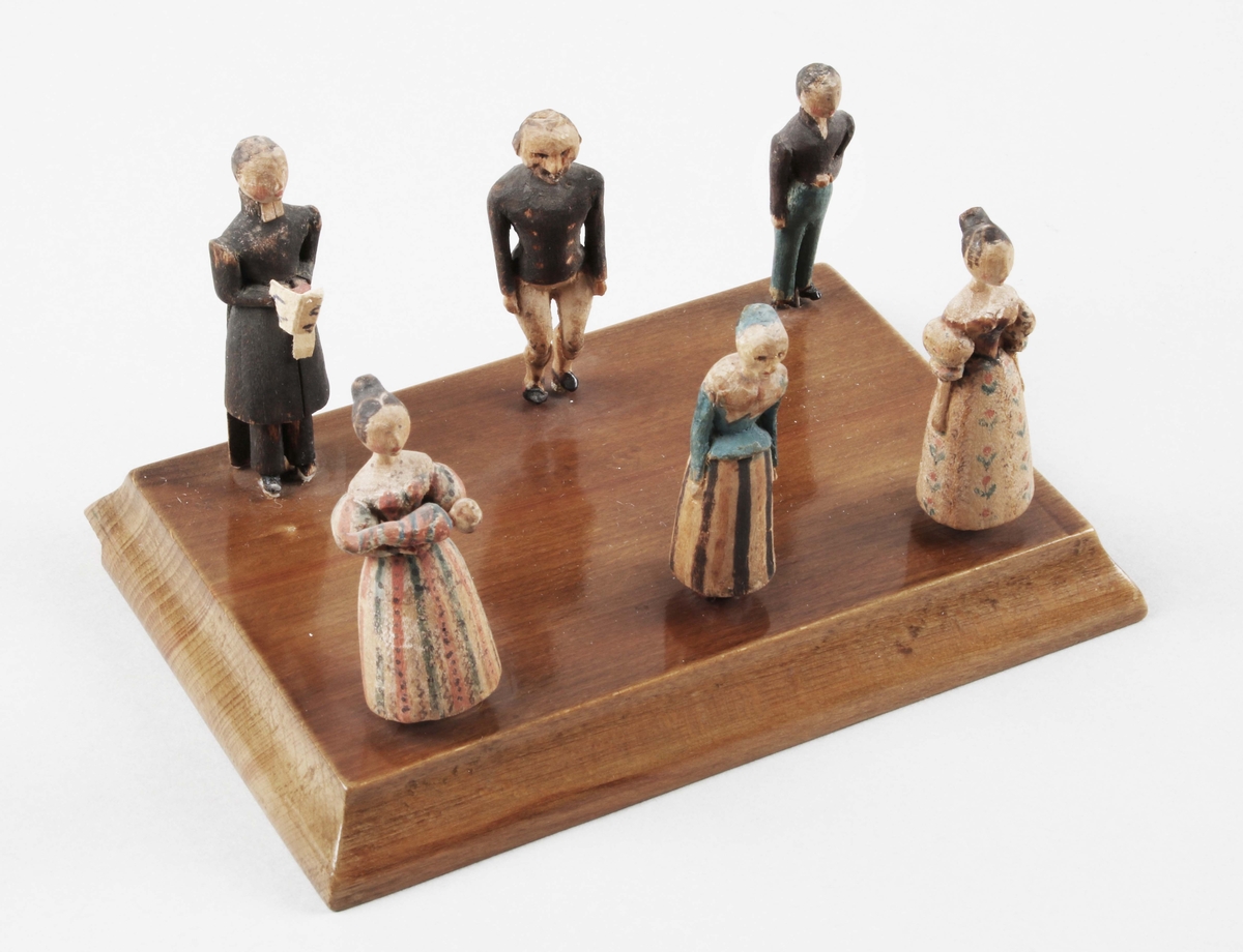 Figurgrupp i trä. "Symboliserar ett dop". 1830-tal. 6 stående figurer, i snidat trä, med handmålad dekor. I form av, präst, kvinna med barn i famnen, 2 kvinnor och 2 män. De sitter med stift, i rektangulär, brunbetsad- och lackad träplatta, med insvängda kanter.

Till denna figurgrupp hör konsolhylla BM 39798.